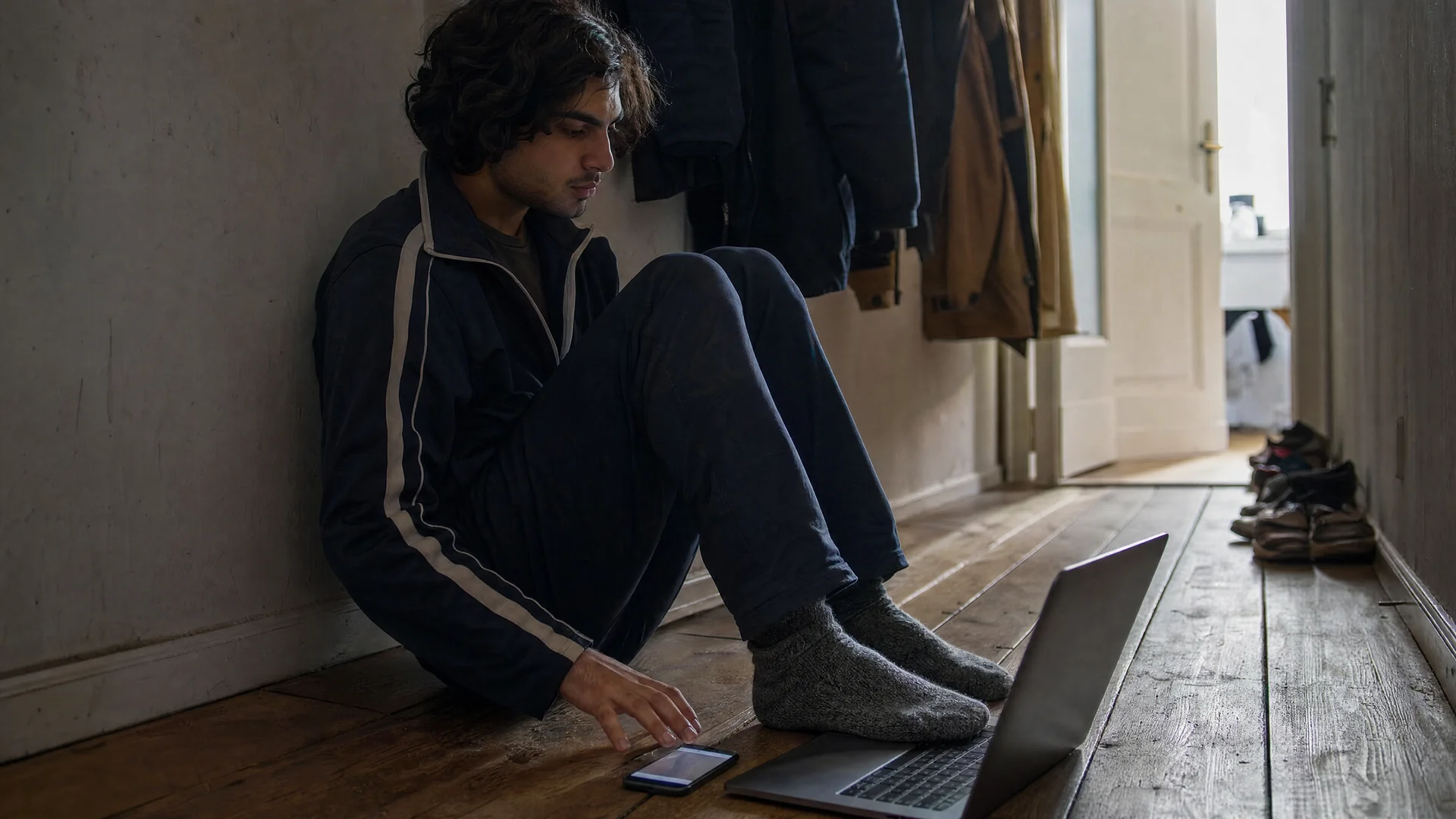 Student sitzt im Flur einer Wohngemeinschaft auf dem Boden, den Laptop vor sich, und spult auf dem Smartphone ein Tutorial-Video zurück.