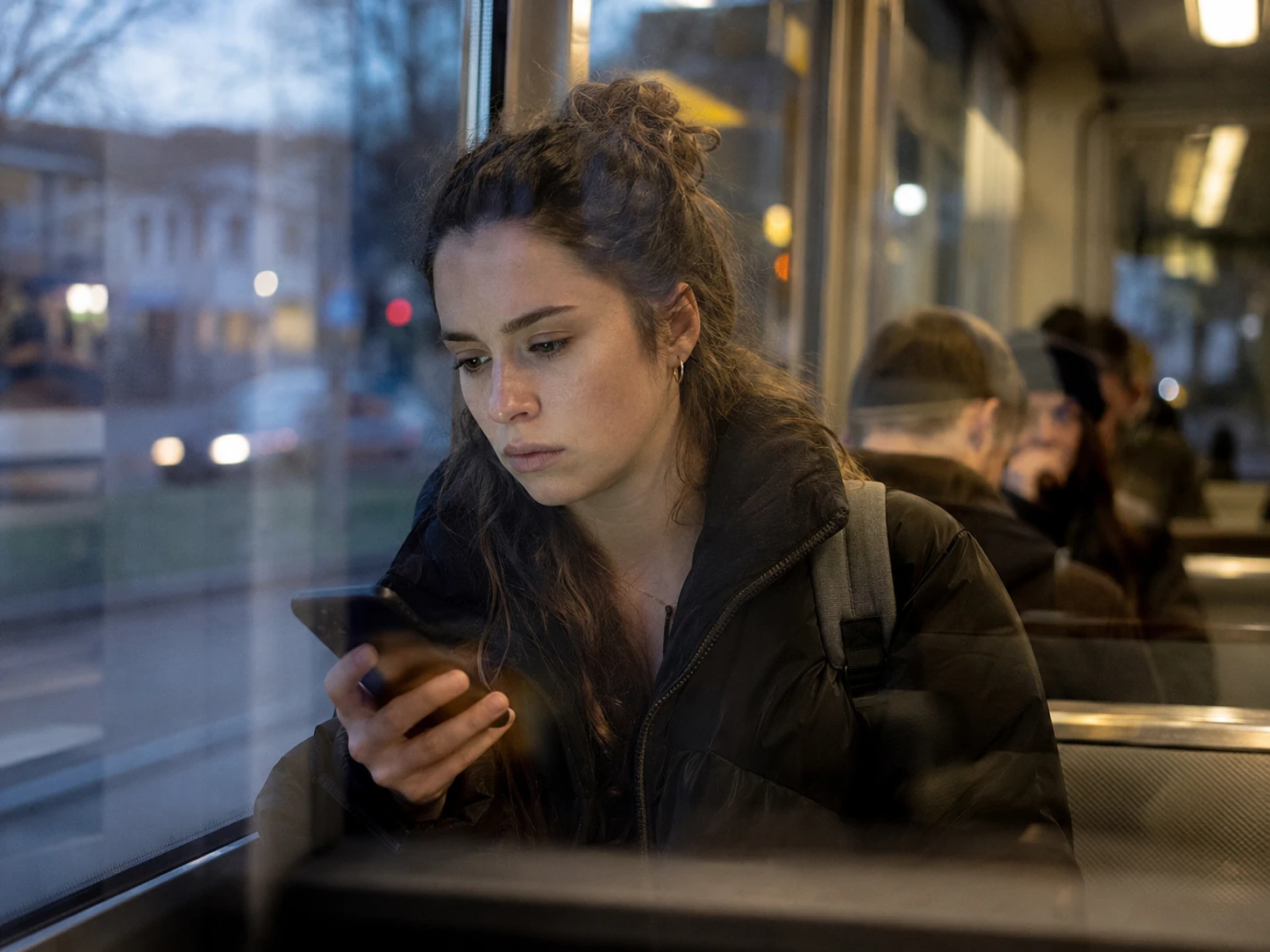 Studentin sitzt in einer Straßenbahn und blickt verunsichert auf ihr Smartphone.