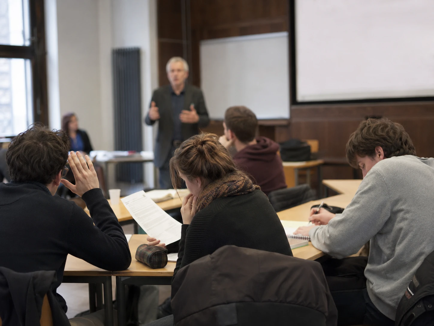 Dozent erläutert einen Sachverhalt vor mehreren zuhörenden, teils verunsicherten Studierenden.