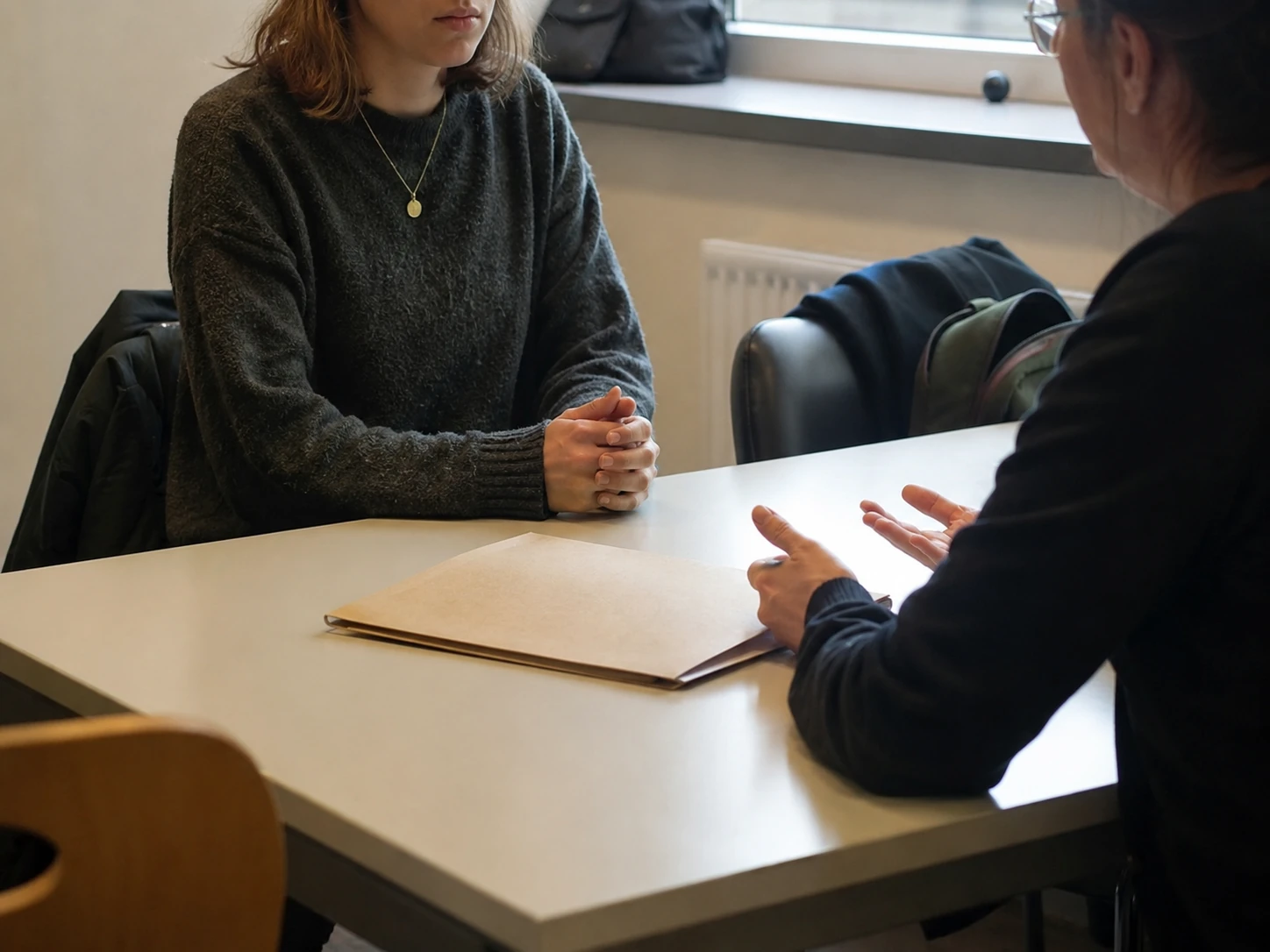 Studentin sitzt an einem Tisch einer ruhigen Beratung gegenüber, die Hände gefaltet und zuhörend, während die beratende Person mit der Hand gestikuliert, zwischen beiden eine aufgeschlagene Mappe.