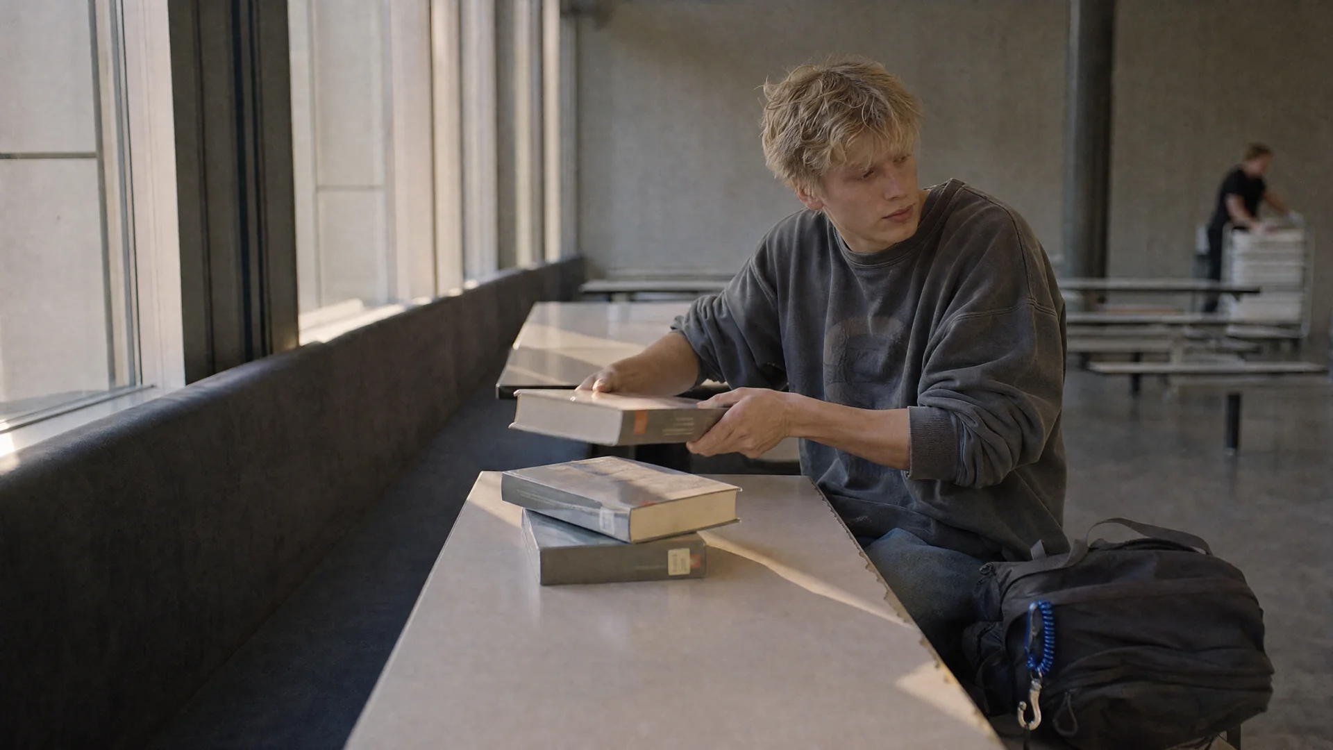 Student sitzt seitlich an einem Mensatisch im flachen Nachmittagslicht und hebt einen von drei Bibliotheksbänden mit beiden Händen vom Tisch auf die Bank neben sich.